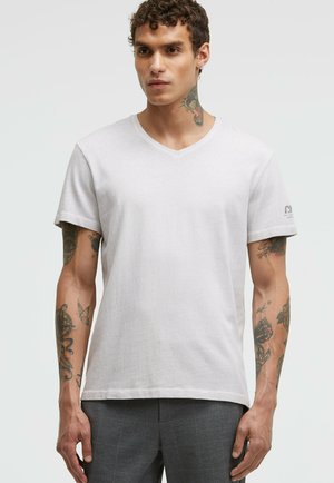Light gray v-neck t-shirt made from soft fabric, featuring short sleeves and a subtle logo on the left sleeve, worn with patterned trousers.