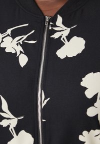 Svart bomberjacka med vitt blommönster, utrustad med dragkedja framtill, ribbad krage och slät tygstruktur.