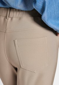 Beige broek van een textuurstof, met een achterzak en een gladde tailleband. Het materiaal heeft een lichte stretch.