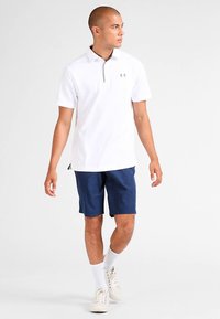 Tânăr care merge, purtând un tricou polo alb cu mâneci scurte și fermoar, pantaloni scurți bleumarin, șosete albe și pantofi sport albi pe un fundal deschis.