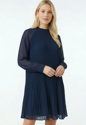 Femme aux longs cheveux blonds portant une robe plissée bleu marine à manches longues transparentes, debout sur un fond blanc.