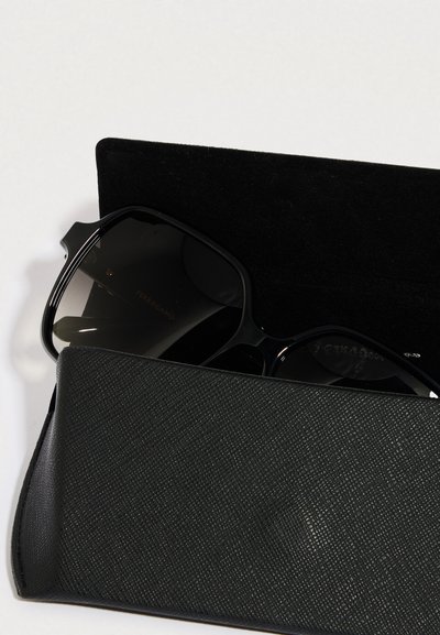 Svarte solbriller med mørke glass delvis synlige inne i et teksturert svart etui. Logo preget på solbrillene. Etuiet har et slankt design.
