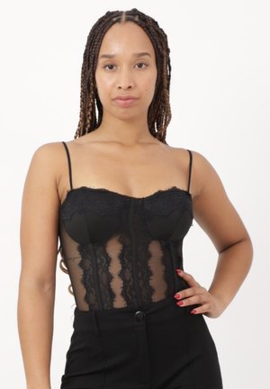 Vrouw met gevlochten haar, gekleed in een zwarte kanten korset-top en zwarte broek, poseert met één hand op de heup tegen een effen achtergrond.
