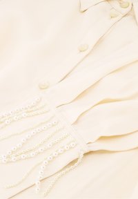 Crème blouse met een voorpand met knopen, gerimpeld ontwerp en decoratieve strengen witte pioenroos aan de onderrand.