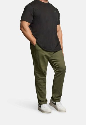 Mann trägt ein schwarzes Kurzarm-T-Shirt, olivgrüne Hose und weiße Sneaker mit schwarzem Swoosh, steht mit den Händen in den Taschen.