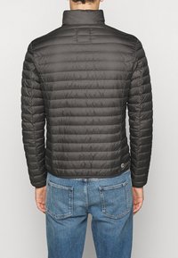 Svart, vadderad pufferjacka med vertikala sömmar, hög krage och elastiska ärmslut. Har en logotyp på vänster nederkant.