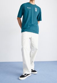 Camiseta de algodón en color teal con logo gráfico y letras en blanco, combinada con pantalones de pierna ancha blancos y zapatillas en blanco y negro sobre un suelo azul.