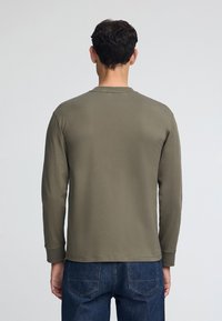 Olivgrön tröja med långa ärmar i mjukt tyg, med rund hals och rak fåll, bärs med jeans, sedd bakifrån.