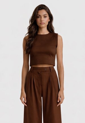 Donna con lunghi capelli castani che indossa un top corto marrone senza maniche e pantaloni marroni a vita alta e gamba larga, in piedi davanti a uno sfondo semplice.