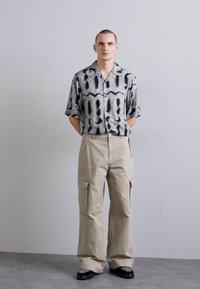 Chemise à manches courtes avec un motif abstrait noir et gris, associée à un pantalon cargo beige avec poches et une coupe décontractée.