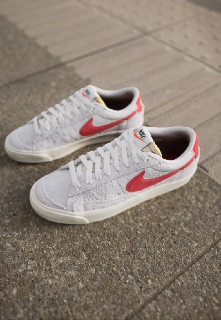 Sneakers bianchi con una finitura texture e accenti rossi. Le caratteristiche includono lacci bianchi, un'etichetta gialla e un design a profilo basso.