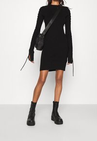 Robe en maille noire à manches longues, découpes aux poignets, et coupe ajustée. Associée à des bottines de combat noires à lacets et un petit sac à bandoulière.