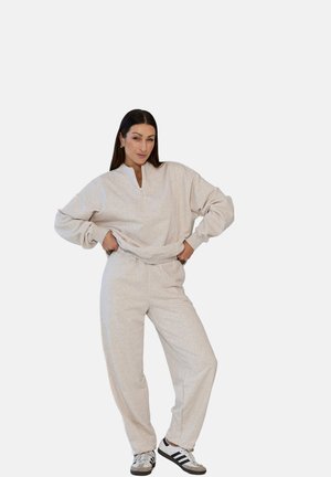 Kobieta pozująca z rękami na biodrach, ubrana w jasnoszary, oversize'owy sweter i dopasowane spodnie dresowe oraz białe i czarne sneakersy.