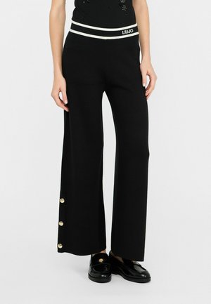 Zwarte wijde broek met een wit gestreepte tailleband, voorzien van gouden knopenaccenten aan de zijkant. De stof heeft een gladde textuur.