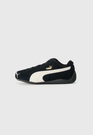 Baskets Puma noires avec bande blanche et logo Puma doré, design profil bas et semelle noire texturée sur fond gris clair.