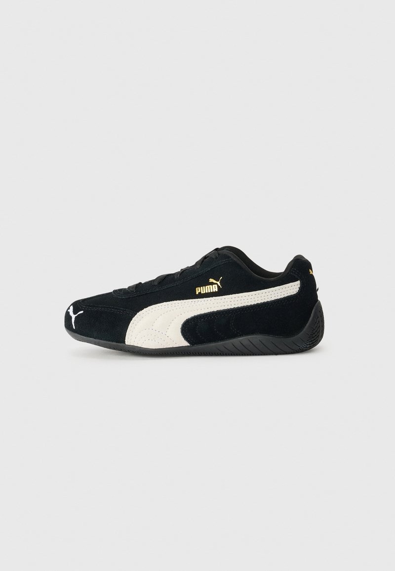 Baskets Puma noires avec bande blanche et logo Puma doré, design profil bas et semelle noire texturée sur fond gris clair.