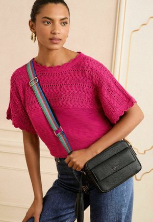 Donna che indossa un luminoso top lavorato a maglia rosa e jeans, con una borsa a tracolla nera testurizzata con una tracolla a strisce arcobaleno e orecchini dorati.