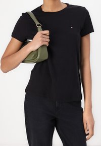 Czarny bawełniany t-shirt z okrągłym dekoltem, z małym logo. Modelka trzyma małą oliwkowozieloną skórzaną torebkę na pasku. Ciemne dżinsy dopełniają stylizację.