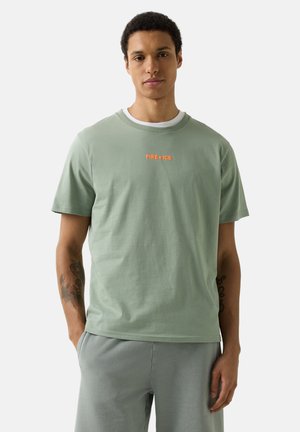 Young man with tattoos wearing a light green "FIRE + ICE" T-shirt and gray pants, standing with hands in pockets against plain background.