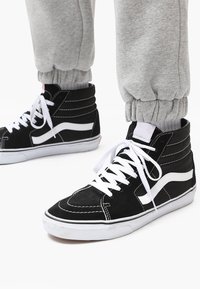 Black high-top sneakers with white accents, featuring suede material, white laces, and a textured design. Pants have elastic cuffs.