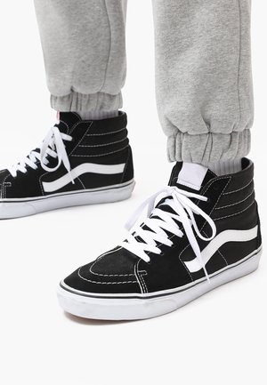 Black high-top sneakers with white accents, featuring suede material, white laces, and a textured design. Pants have elastic cuffs.