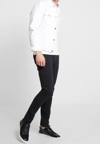 White denim jacket with dark buttons, black slim-fit pants, and black sneakers with white soles, showcasing a modern casual style.