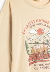 Beżowa bawełniana koszulka z krótkim rękawem, z okrągłym dekoltem, ozdobiona graficznym nadrukiem gór, niedźwiedzia oraz napisem "Największa Natura".