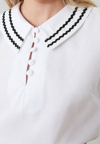 Valkean paidan, jossa on terävä kaulus mustalla sahalaitatrimmin ja nappisuljin, jossa on pyöreät valkoiset napit.