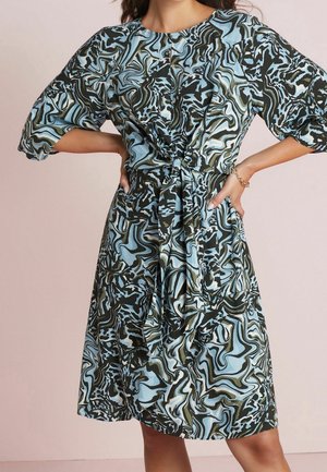 Kvinde iført knælængde kjole med abstrakt mønster i blå, sort og grønne farver, trekvart ærmer og knudedetalje foran i taljen.