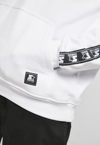 Hanorac alb cu accente negre. Dispune de o buzunar, manșete ribbed și un patch dreptunghiular negru cu un design în formă de stea.