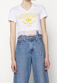 Vit kortärmad croptop med gul "Adidas Athletic Club" grafik. Spetsdetalj vid fållen. Kombinerad med högmidjade ljusblå jeans.