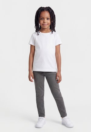 Κορίτσι με πλεξούδες, που φοράει λευκό κοντομάνικο πουκάμισο, γκρι τζιν και λευκά αθλητικά παπούτσια, στέκεται μπροστά σε μια απλή ταπετσαρία.