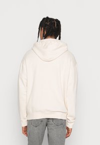 Světlý béžový mikina s volným střihem, strukturovanou látkou, kapucí se stahovací šňůrkou a žebrovanými manžetami. Doplňena světle šedými džínami.