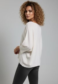 Weißer Oversize-Pullover mit dreiviertel Ärmeln, weicher Strickstruktur und lässiger Passform. Kombiniert mit dunkelgrauen Jeans.