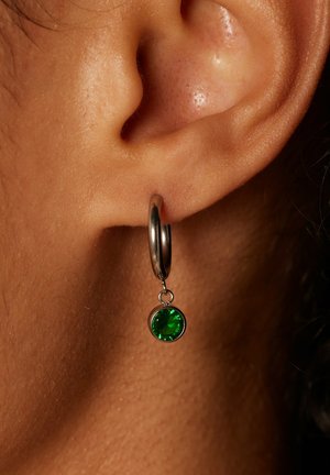 Zilveren oorring met een groene edelsteen hanger, tegen de huid. De oorring heeft een gladde textuur en een ronde vorm.
