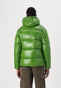 Zeleni puffer jakna s sjajnom završnom obradom, šivenim dijelovima i kapuljačom. Ima elastične manžete i bočne džepove, u kombinaciji s maslinastim cargo hlačama.