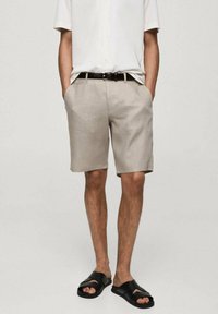 Pantalones cortos de lino beige con bolsillos laterales, frente plano y un corte relajado. Se combinan con una camisa blanca de manga corta y sandalias negras.