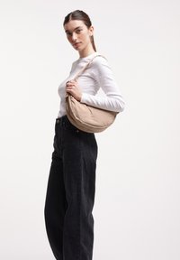 Borsa a spalla in tessuto beige e morbido, con una forma arrotondata, dotata di una tracolla corta e di dettagli di cucitura discreti. Indossata sulla spalla.