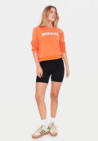 Oranje sweatshirt met "BISOU" in witte tekst, gecombineerd met zwarte fietsbroekjes. Model draagt beige sneakers met groene accenten en witte sokken.