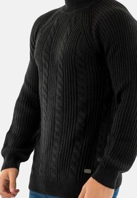 Maglione nero a trecce con collo alto, caratterizzato da coste testurizzate e un piccolo logo metallico vicino all'anca.