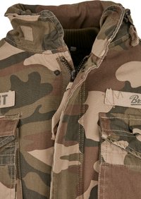 Maskáčová bunda v béžových a zelených odtieňoch s vysokým golierom, zipsovým zapínaním a dvoma náprsními vreckami; má robustnú tkaninu a vojenský dizajn.