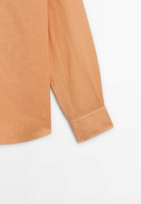 Manche et poignet d'une chemise à manches longues marron clair en tissu texturé sur fond blanc.