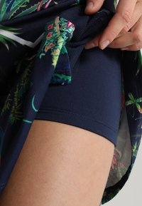 Pantaloni corti in tessuto a motivo floreale di un blu navy scuro con unafodera in rete. Dettaglio della stampa floreale e del risvolto dei pantaloni corti solidi con una mano che regola il tessuto.
