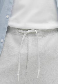 Lys grå sweatpants med en blød tekstur, der har en snorelukning i hvid og en ribbet talje. En blåstribet skjorte med knaplukning ovenfor.