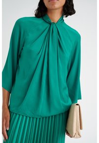 Blusa verde acqua con un collo alto e tessuto arricciato. Le maniche sono ampie e la modella tiene in mano una borsa a tracolla trapuntata beige.