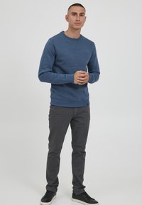 Kék pamut pulóver bordás mandzsettával és alsó peremmel, szürke, slim-fit nadrággal és fekete sportcipővel párosítva, fehér háttér előtt állva.