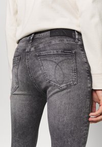 Jeans de mezclilla gris con un ligero desgaste, que cuentan con un diseño clásico de cinco bolsillos, una etiqueta de cuero negro con el logo y detalles de costura distintivos.