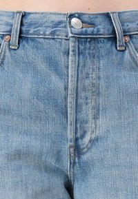 Κοντινό πλάνο στη μέση από ανοιχτό μπλε τζιν παντελόνι, που δείχνει το κουμπί, τις θηλιές της ζώνης και τις λεπτομέρειες ραψίματος των μπροστινών τσεπών.