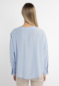 Una persona con capelli lunghi indossa una blusa azzurro chiaro, girata di spalle, su uno sfondo bianco semplice. La blusa ha una vestibilità comoda e polsini con bottoni.