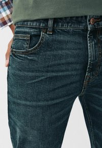 Tummansiniset farkut kapealla istuvuudella, joissa on normaali taskumalli, vastavärinen oranssi ompelu ja metallinen napin suljin.